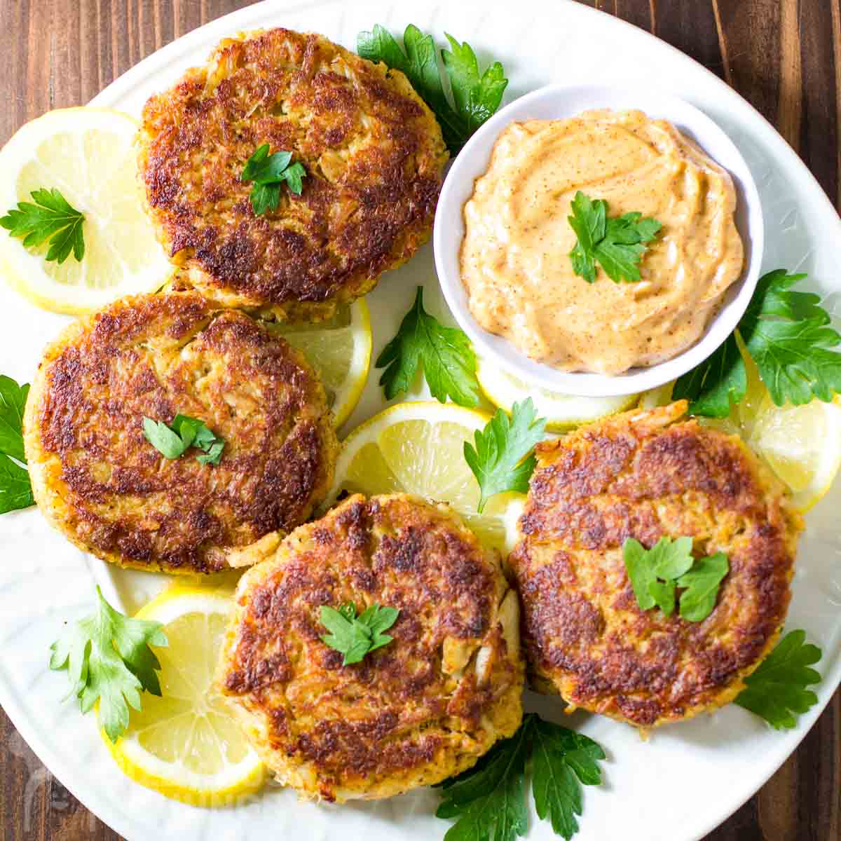 Overhead shot of 4 crab cakes on a bed of lemon slices garnished with fresh herbs with a bowl of remoulade on the side all on a white plate.