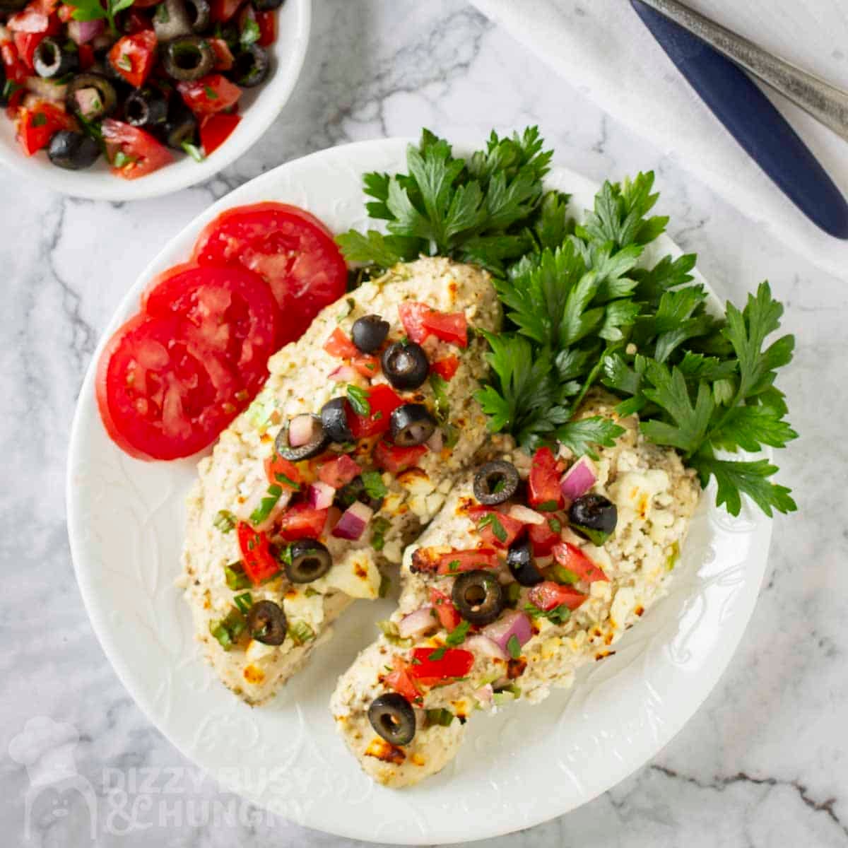 Overhead shot of two mediteranean chicken breasts with fresh cilantro and tomato slices on the side on a white plate.