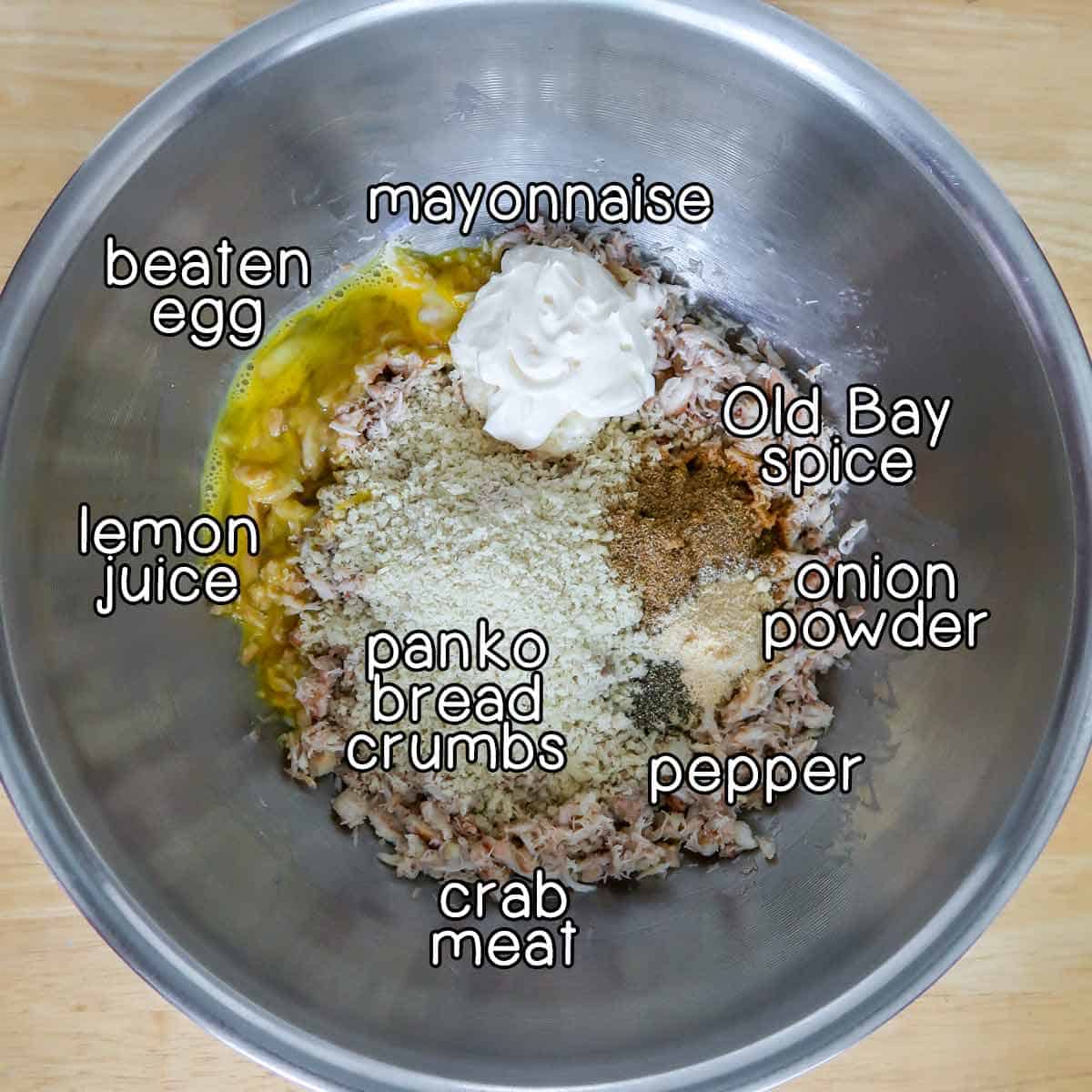 Overhead shot of ingredients - mayonnaise, beaten egg, lemon juice, panko bread crumbs, crab meat, old bay spice, onion powder, and pepper.