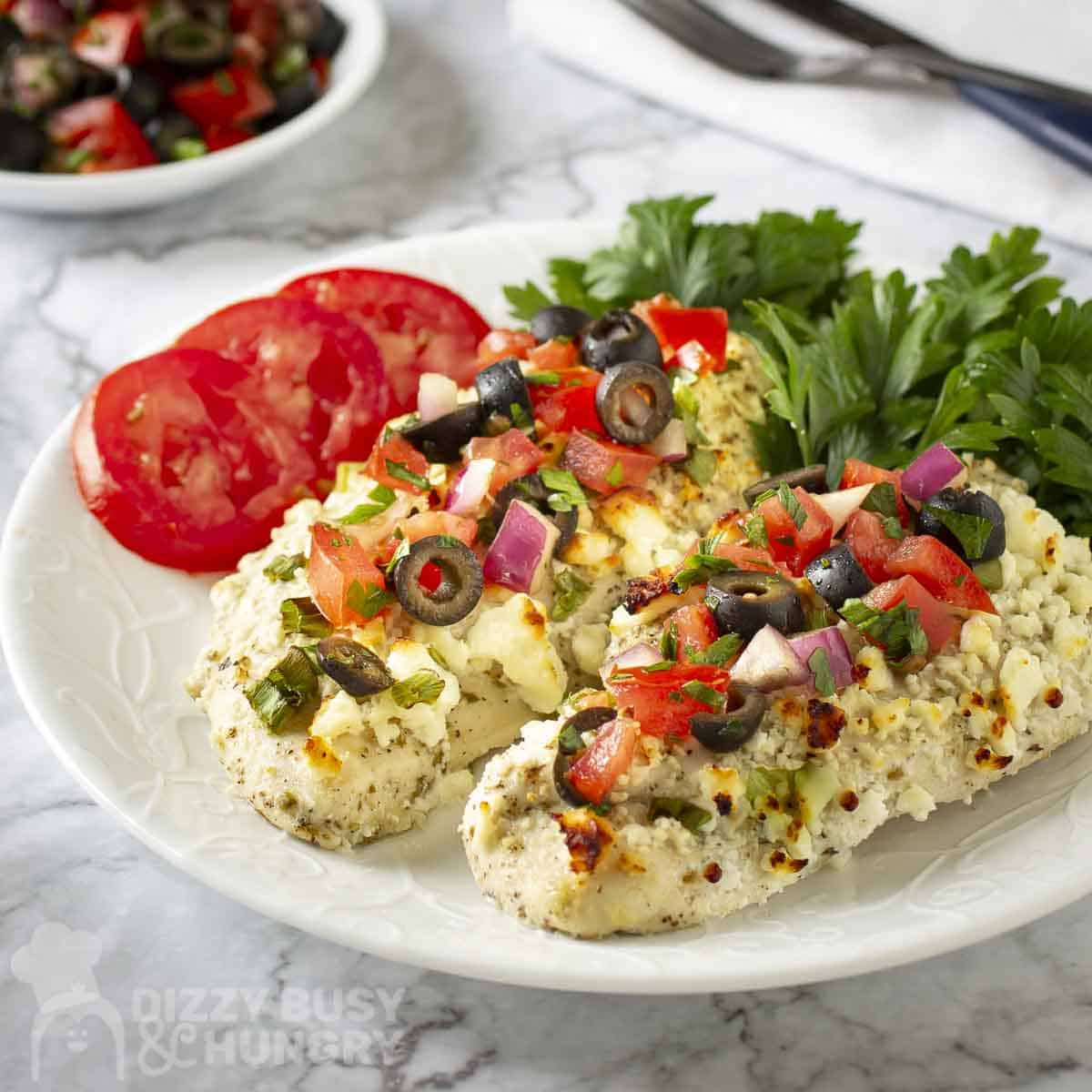 Side shot of two mediteranean chicken breasts with fresh cilantro and tomato slices on the side on a white plate.