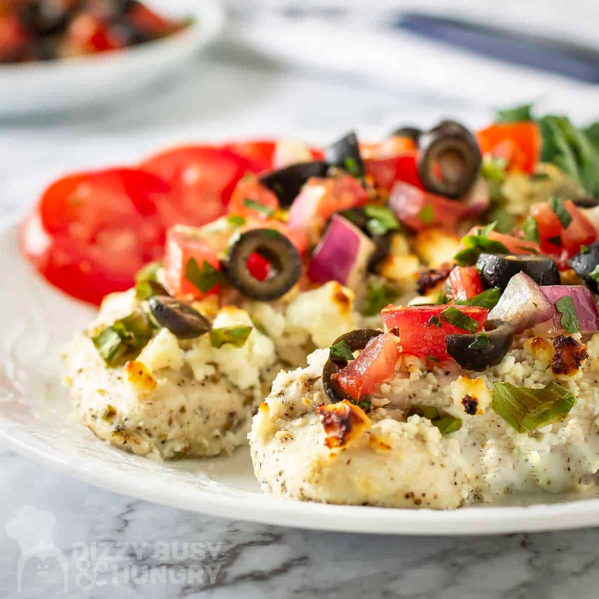 Side close up shot of two mediteranean chicken breasts with fresh cilantro and tomato slices on the side on a white plate.