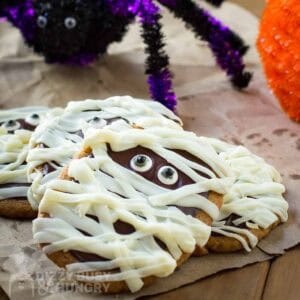 Side shot of mummy cookies laying on a brown paper towel with fake spider and pumpkin halloween decorations in the background.