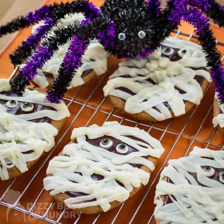 Overhead close up shot of mummy cookies on a wire cooling rack with an burnt orange surface underneath and a fake spider decoration on top of the cookies.