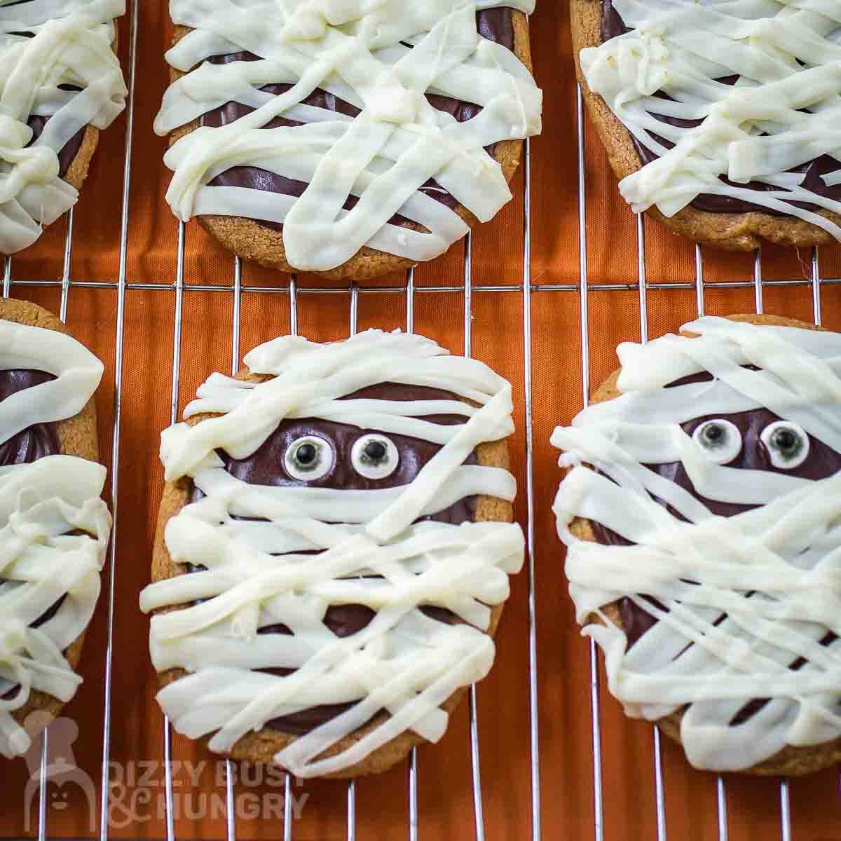 Overhead close up shot of mummy cookies on a wire cooling rack with a burnt orange surface underneath.