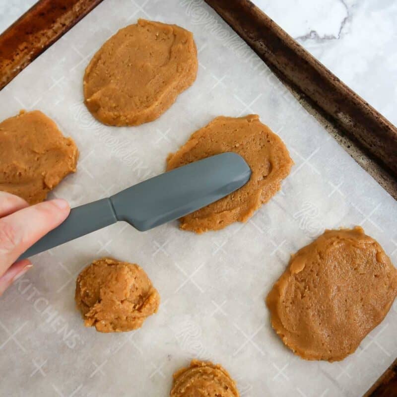 Overhead view of process shot 2 - line a baking sheet with parchment paper, then use a cookie scoop to scoop out the dough and place on the parchment paper. With the back of a spoon or spatula, flatten the balls of dough and shape them into rectangular ovals.