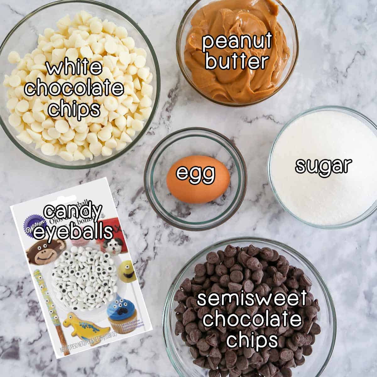 Overhead shot of ingredients - white chocolate chips, peanut butter, egg, sugar, semisweet chocolate chips, and candy eyeballs.