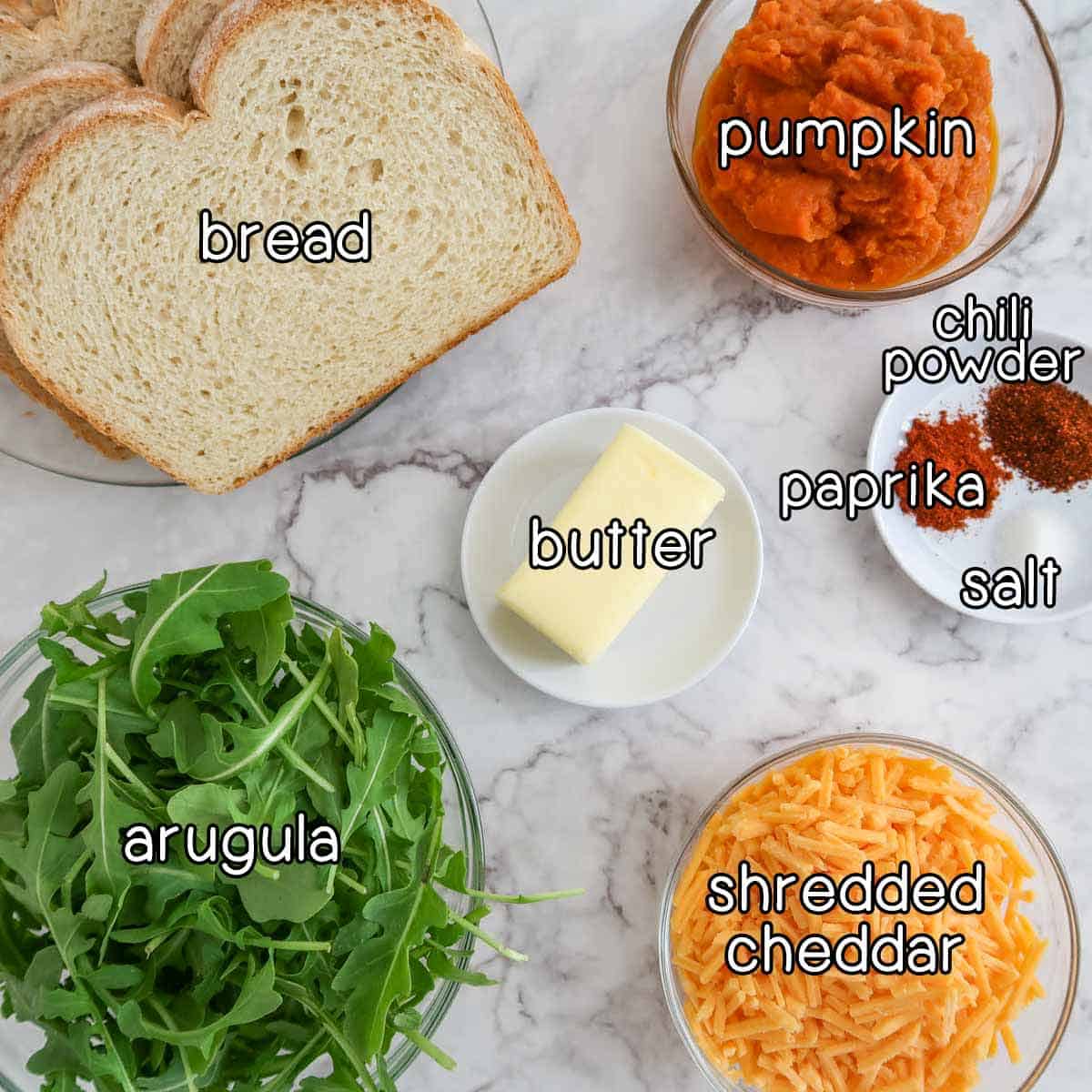 Overhead shot of ingredients - bread, pumpkin, arugula, shredded cheddar, butter, chili powder, paprika, and salt.