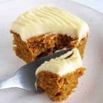 Side shot of a fork taking a piece of pumpkin spice cake with cream cheese frosting off of a white speckled plate.