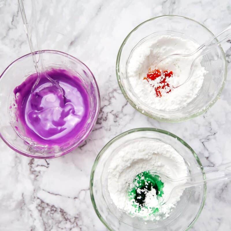 Overhead view of process shot 3 - Add ½ cup powdered sugar and ½ tablespoon water in each of 3 small bowls. Add a few drops of food coloring in each to create the orange, green, and purple colors. Mix well until the icing is stiff. Add more food coloring, a drop or two at a time, until the colors are vibrant enough for you.