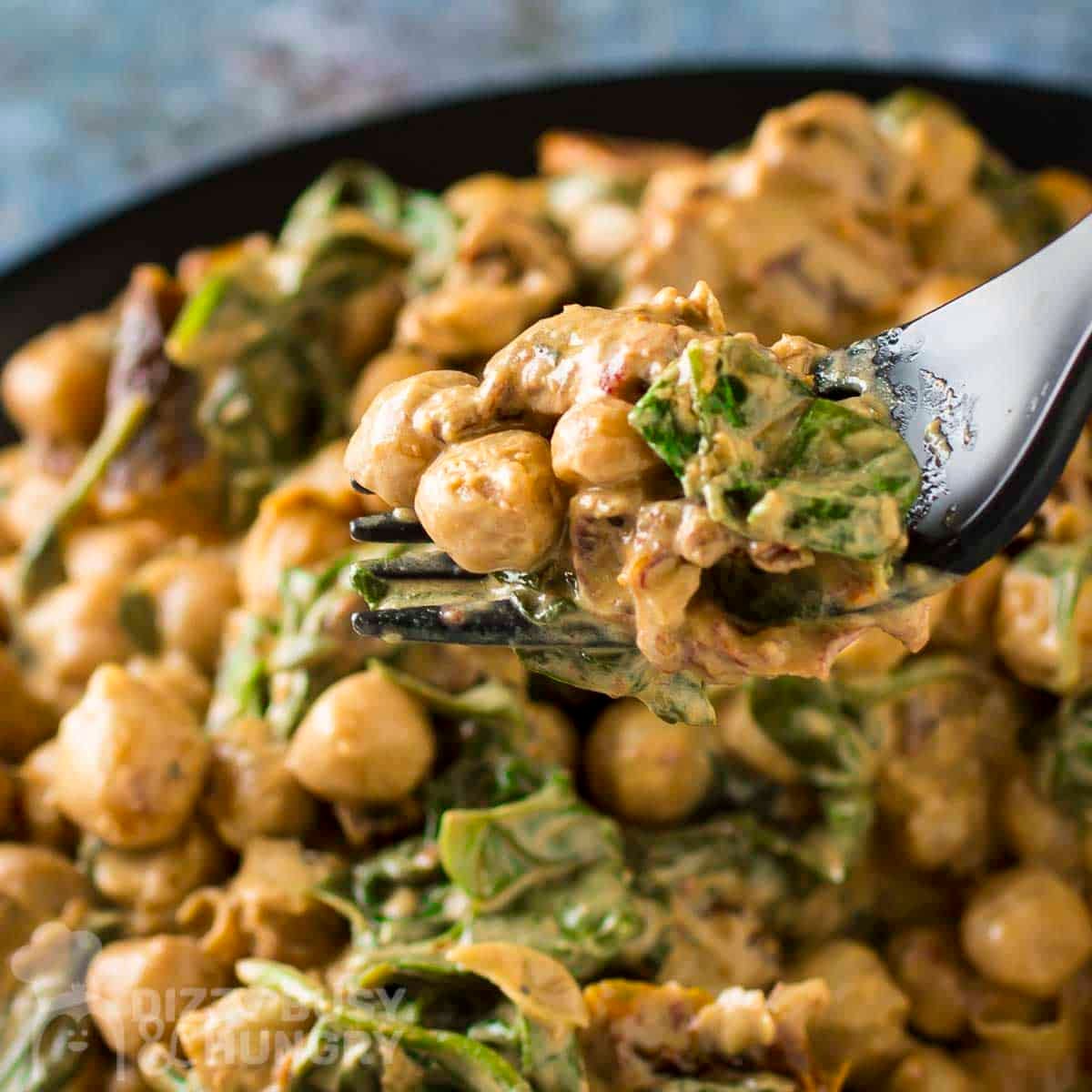 Close up shot of a fork holding up a bite of marry me chickpeas and spinach over a full black plate of the meal.