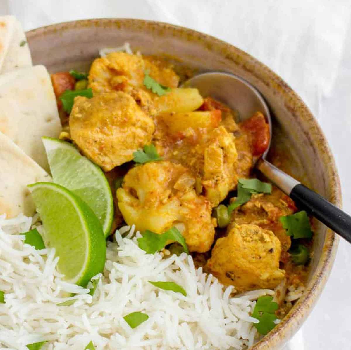 Close up shot of chicken curry with rice and pita bread slices in a brown speckled bowl garnished with fresh parsley and two lime wedges with a spoon on the side.