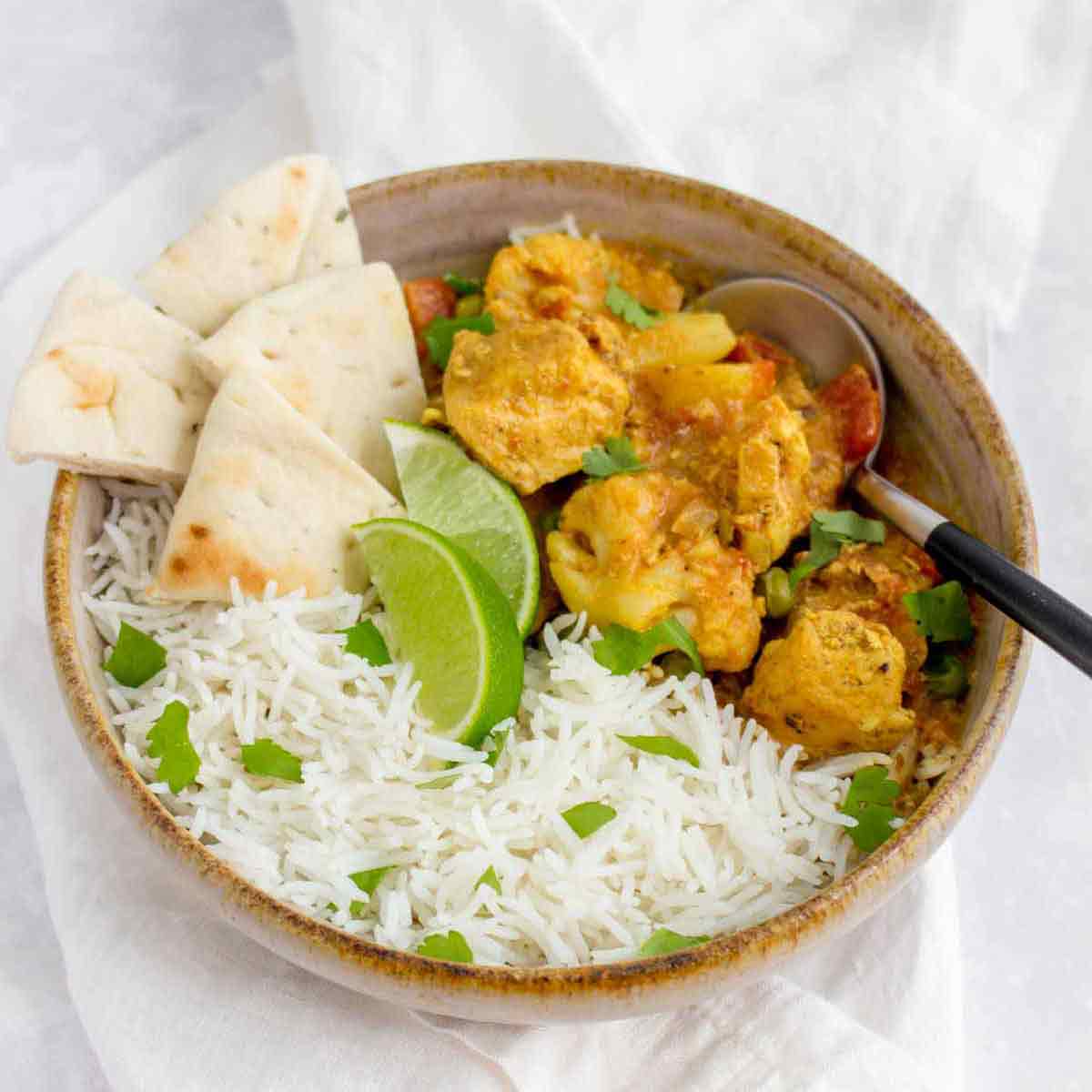 Side angled shot of chicken curry with rice and pita bread slices in a brown speckled bowl garnished with fresh parsley and two lime wedges with a spoon on the side.