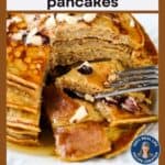 Side shot of a fork taking a piece out of a stack of pumpkin pancakes drizzled with maple syrup and sprinkled with chopped walnuts.