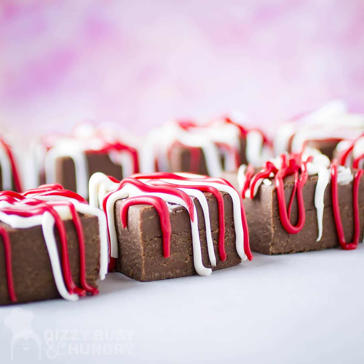 Side shot of multiple pieces of fudge drizzled with red and white melted chocolate on a white surface.